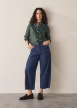 Woman wearing dark green ruffle front shirt with blue jeans and loafers, set against a beige background
