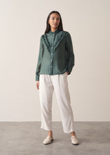Woman wearing a dark green blouse with ruffled details and cream suit trousers.