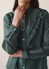 Woman wearing a dark green blouse with ruffled details and cream suit trousers.