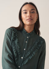 Woman wearing a dark green blouse with ruffled details and cream suit trousers.