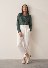 Woman wearing a dark green blouse with ruffled details and cream suit trousers.