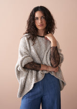 Woman wearing a black lace top with beige jumper and blue jeans, against a beige background