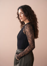 Woman wearing a black lace top with sheer sleeves, paired with green satin trousers, against a beige background