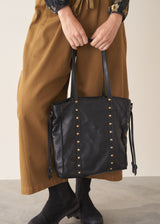 A person holds a large black leather tote bag with gold-tone studs and double shoulder straps against a plain beige studio background.
