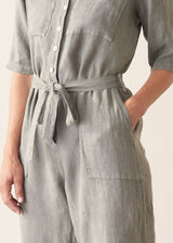 Woman wearing a light sage green linen jumpsuit with a waist tie and pockets