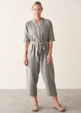 Woman wearing a light sage green linen jumpsuit with a waist tie and pockets