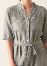 Woman wearing a light sage green linen jumpsuit with a waist tie and pockets