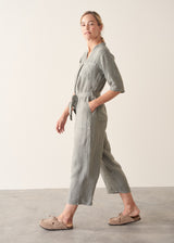 Woman wearing a light sage green linen jumpsuit with a waist tie and pockets