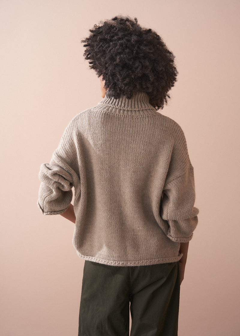 Person wearing a chunky beige fisherman sweater and green barrel leg trousers facing a beige background