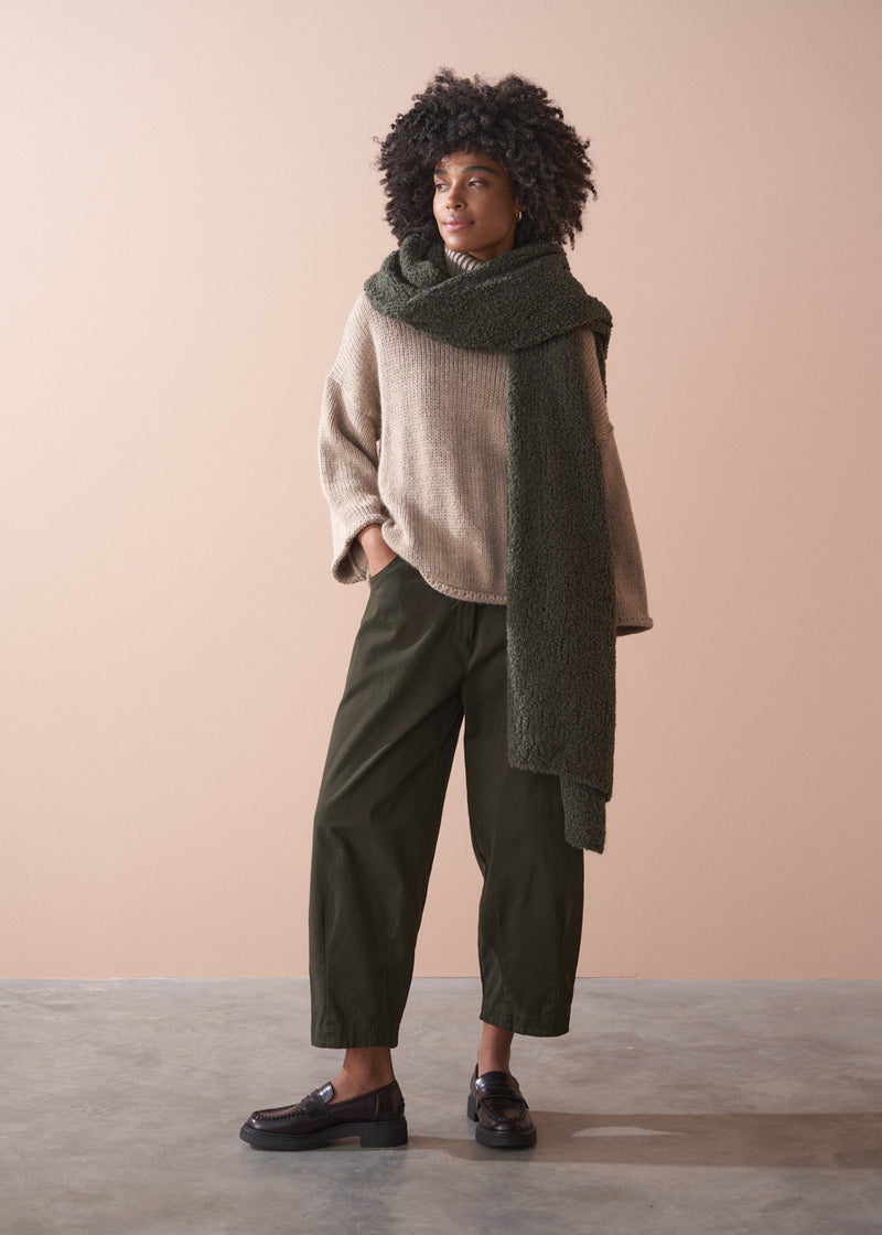 Woman in a chunky knit beige jumper, grey knitted scarf and khaki trousers.