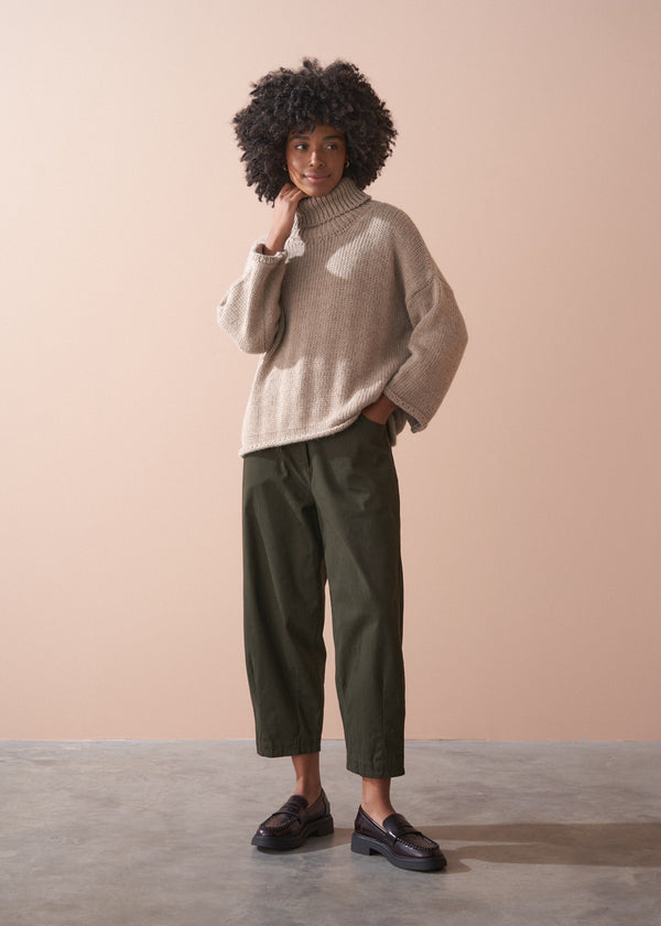 Person wearing a beige roll neck fisherman sweater and green barrel leg trousers against a beige background