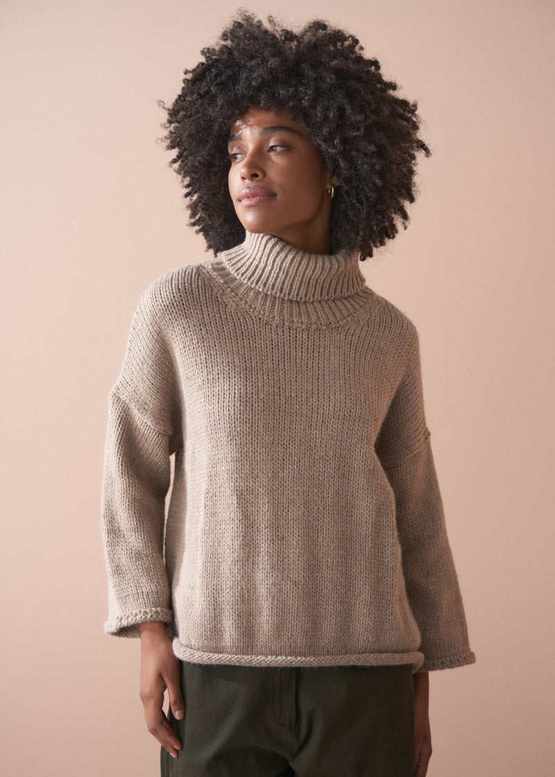 Person wearing a beige roll neck fisherman sweater and green barrel leg trousers against a neutral background