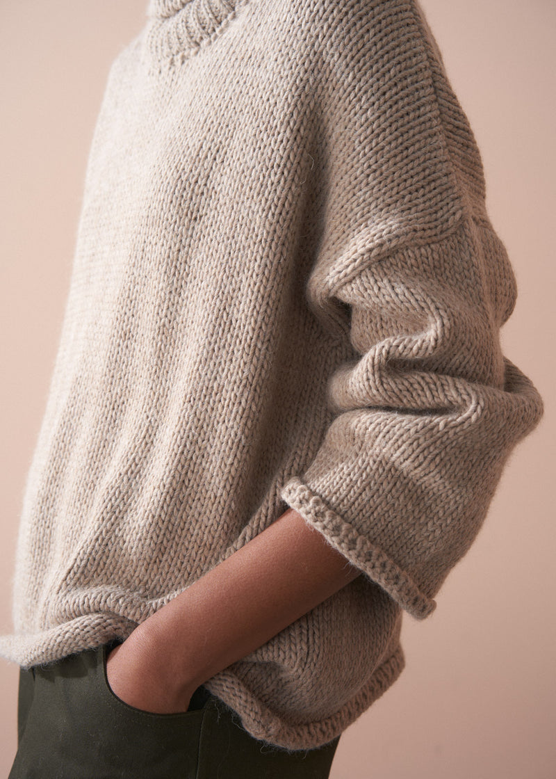 Person wearing a beige fisherman sweater and green barrel leg trousers against a beige background