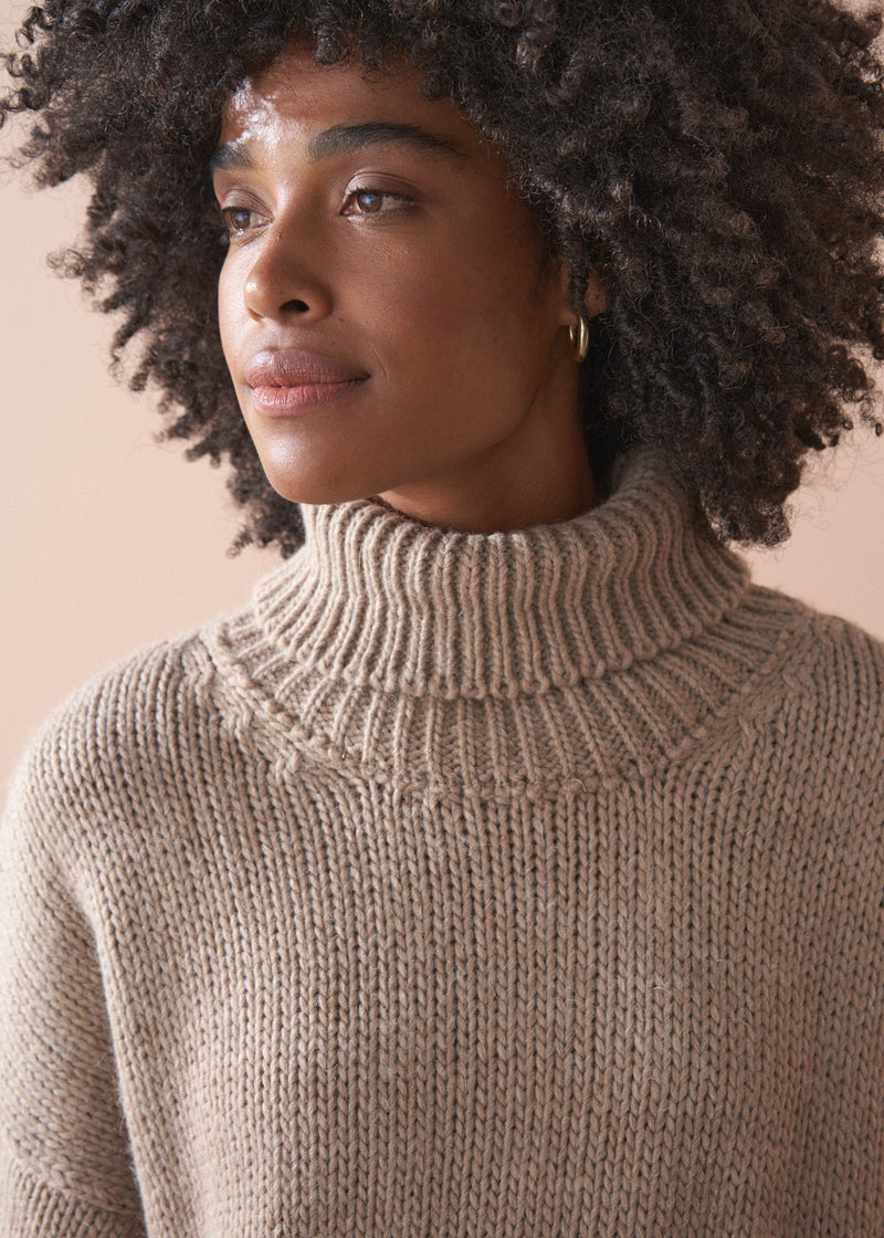 Person wearing a knitted beige fisherman sweater and green barrel leg trousers against a beige background