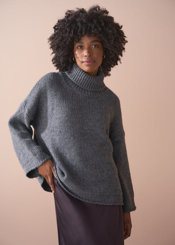 Woman wearing a grey chunky knit sweater and satin bias cut skirt against a beige background