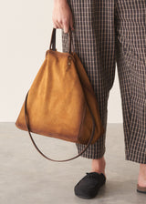 A person wearing brown and black checkered cropped trousers and black slip-on shoes holds a large mustard yellow suede slouchy shoulder bag with a thin brown leather strap against a neutral studio background.