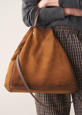 A person wearing a dark gray sweater and brown-and-white checkered trousers carries a large, slouchy tan-brown suede or leather shoulder bag with a thin matching strap.