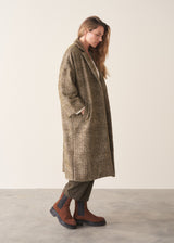 A woman wearing an oversized checked coat with dark green trousers, ginger boots and a white top against a plain background.
