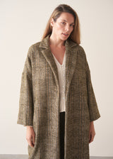 A woman wearing an oversized checked coat with dark green trousers, ginger boots and a white top against a plain background.