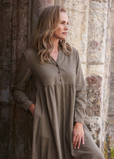 A woman wearing a long-sleeve, button-front midi dress with a small-scale brown and beige gingham pattern, leaning against a rustic stone pillar.