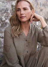 A woman with blonde hair wearing a brown and beige gingham long-sleeve button-up shirt, posing against a textured stone wall.