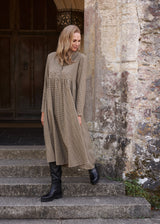 A woman wearing a long-sleeved, knee-length taupe dress and black leather boots stands on stone steps next to a rustic stone wall and wooden door.