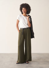 Woman wearing white, round neck t-shirt and dark green wide leg trousers against a plain background