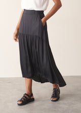 Woman wearing a white top and black satin tiered midi skirt against a plain background