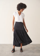 Woman wearing a white top and black satin tiered midi skirt against a plain background