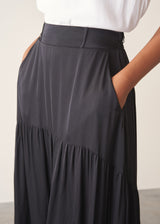 Woman wearing a white top and black satin tiered midi skirt against a plain background