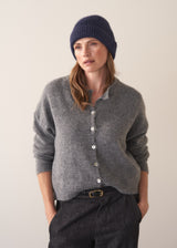 Woman wearing a grey cardigan, black jeans, and a navy wool mix beanie against a plain background
