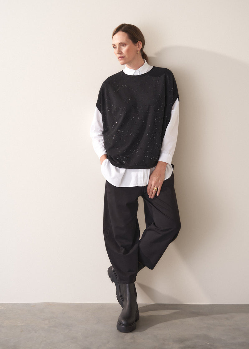 Person wearing a black sweater vest with sequin detail, white shirt, and black trousers, leaning against a plain wall.