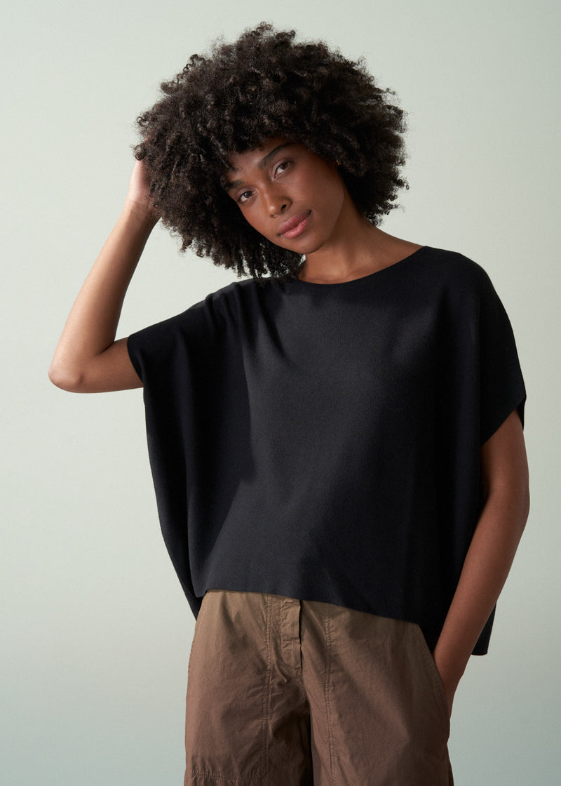 Woman wearing a black sleeveless sweater and brown khaki trousers against a plain background