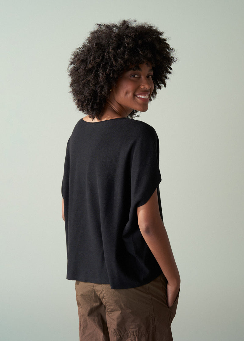 Person wearing a black sleeveless knit and brown pants facing a plain background