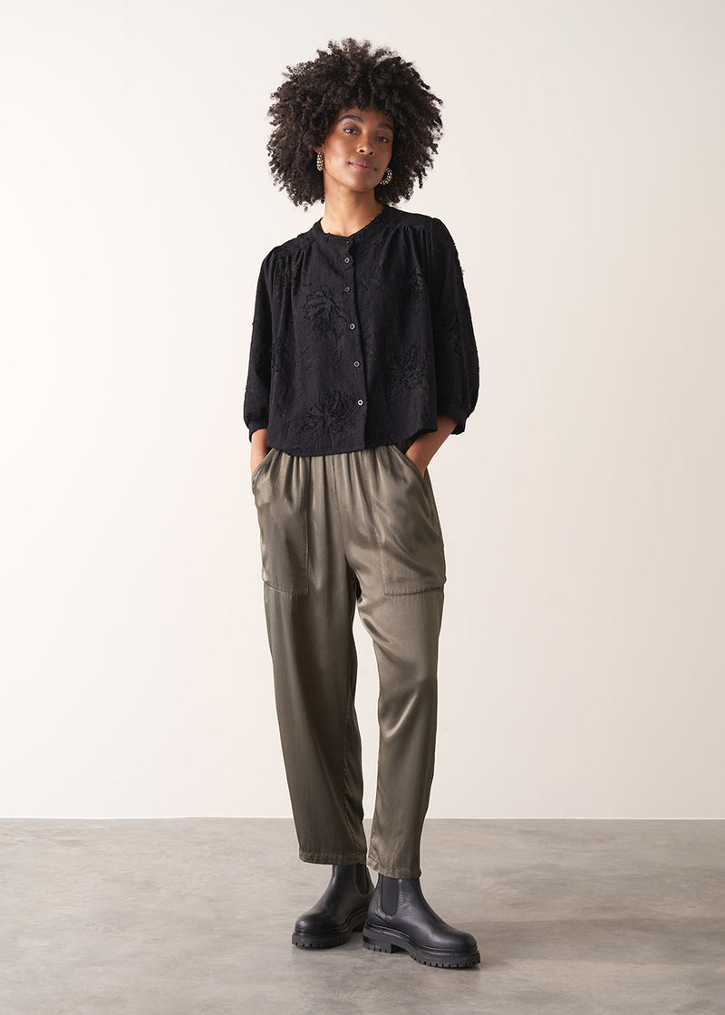 Woman wearing a black vintage-style jacquard blouse with khaki satin trousers on a neutral background