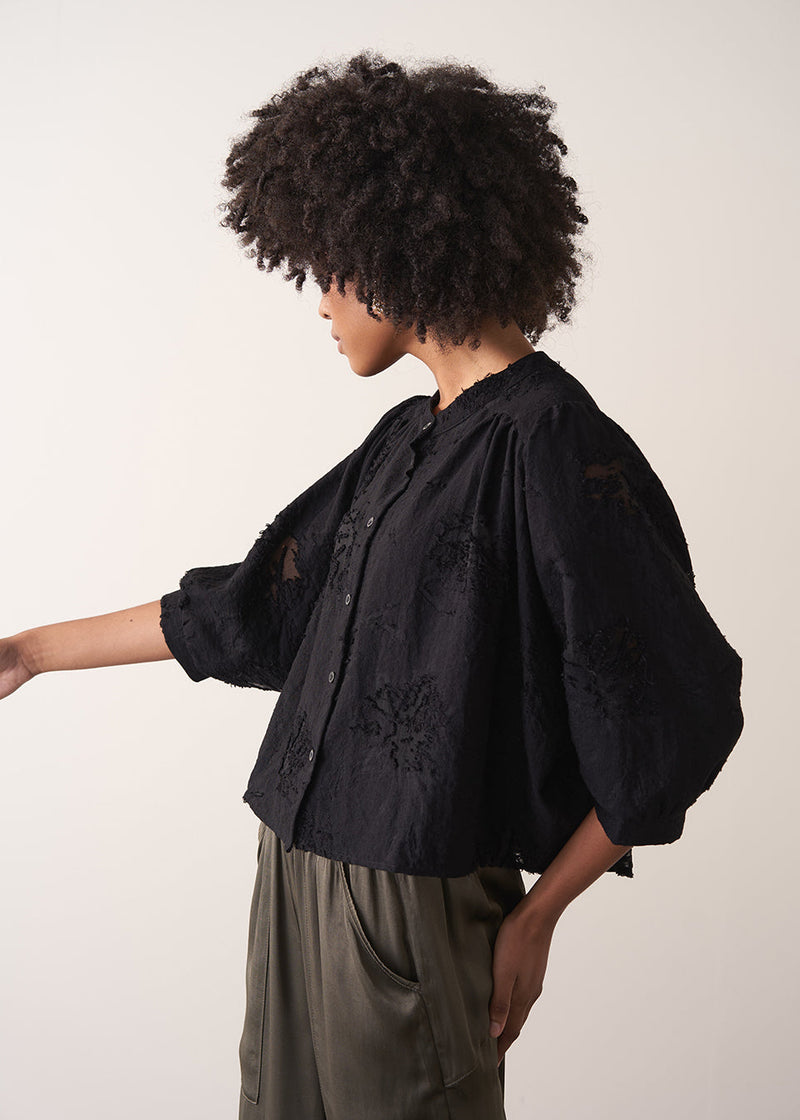 Woman wearing a black vintage-style jacquard blouse with khaki satin trousers on a neutral background