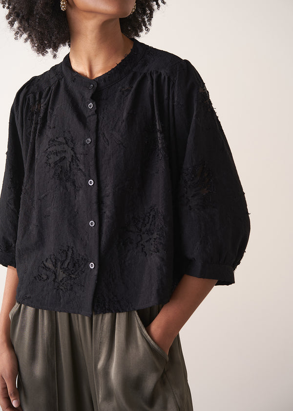 Woman wearing a black vintage-style jacquard blouse with khaki satin trousers on a neutral background