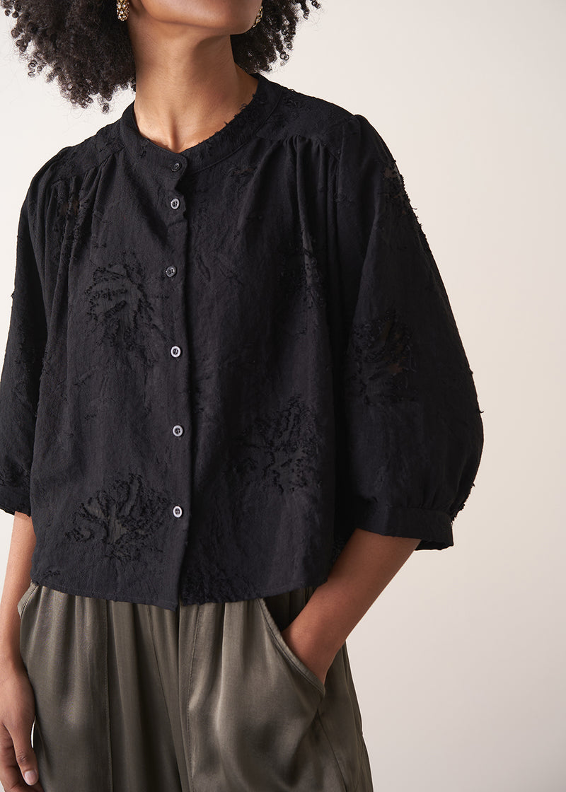 Woman wearing a black vintage-style jacquard blouse with khaki satin trousers on a neutral background