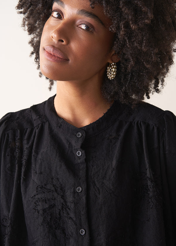 Woman wearing a black vintage-style jacquard blouse with khaki satin trousers on a neutral background