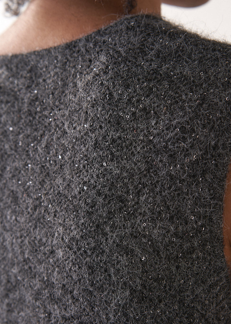 Woman wearing a grey sleeveless sparkle knit sweater and black wide leg satin trousers against a plain background