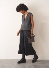 Woman wearing a grey sleeveless sparkle knit sweater and black wide leg satin trousers against a plain background