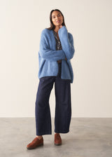 Woman wearing chunky bright blue knit cardigan with patterned blouse and denim trousers against a plain background.