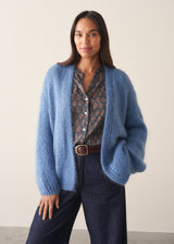 Woman wearing chunky bright blue knit cardigan with patterned blouse and denim trousers against a plain background.