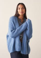 Woman wearing chunky bright blue knit cardigan with patterned blouse and denim trousers against a plain background.