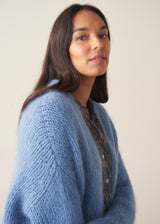 Woman wearing chunky bright blue knit cardigan with patterned blouse and denim trousers against a plain background.