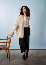 A woman in a soft oatmeal chunky cardigan, white shirt, and black trousers leaning on a wooden chair.
