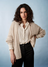A woman in a soft oatmeal chunky knit cardigan, white shirt and black trousers.