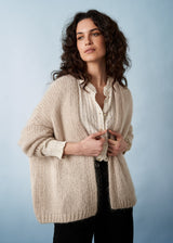 Woman wearing a chunky knit oatmeal cardigan over an off white shirt and black trousers.