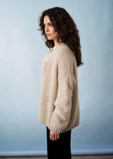 Side view of a woman wearing a chunky knit oatmeal cardigan, over a white shirt and black trousers.