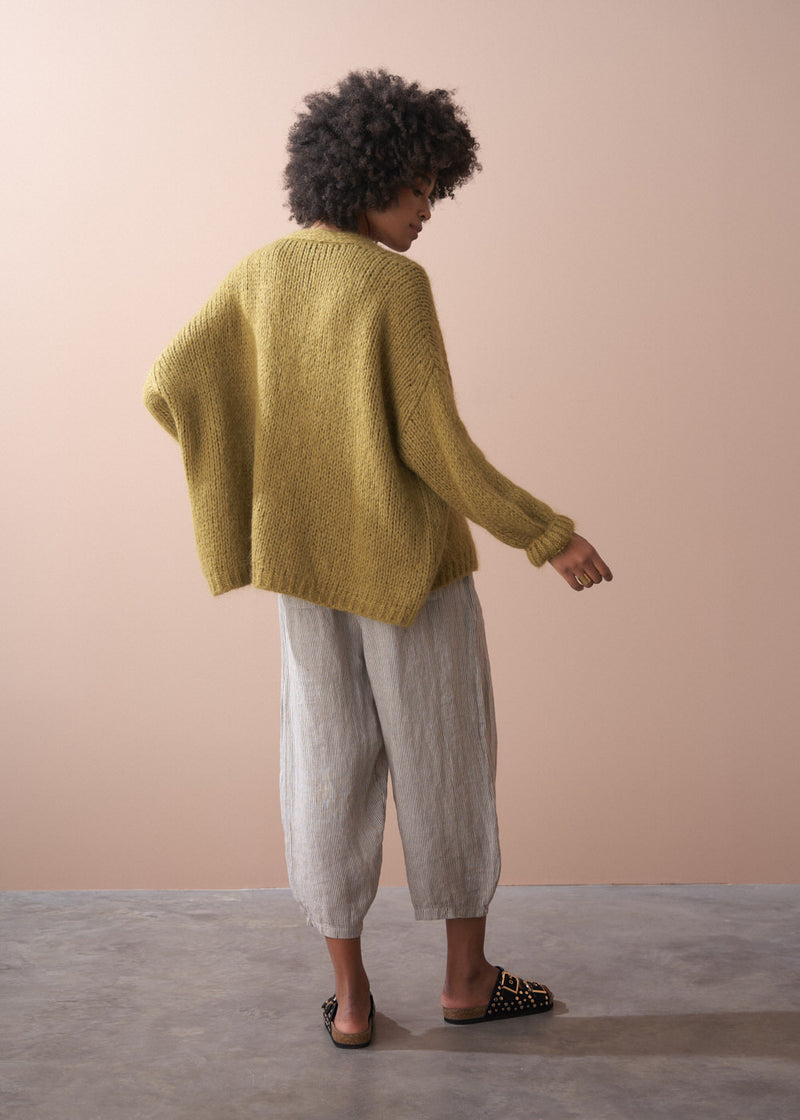 Woman wearing an olive green chunky cardigan and striped trousers, facing a neutral background.